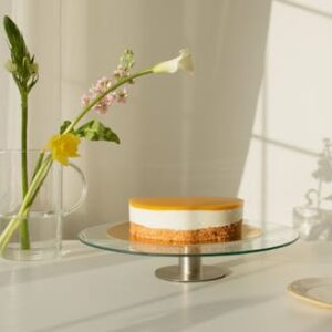 Minimalist lemon cheesecake on a glass stand with fresh spring flowers in vase, bathed in natural light.