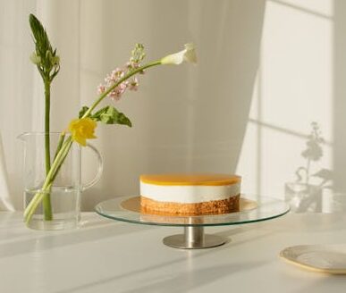 Minimalist lemon cheesecake on a glass stand with fresh spring flowers in vase, bathed in natural light.