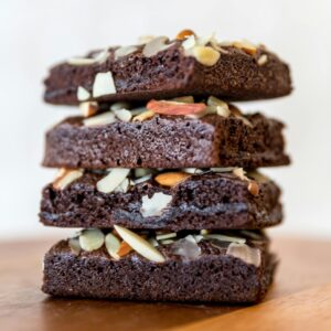 Close-up of stacked chocolate brownies topped with almond slices on wooden surface.