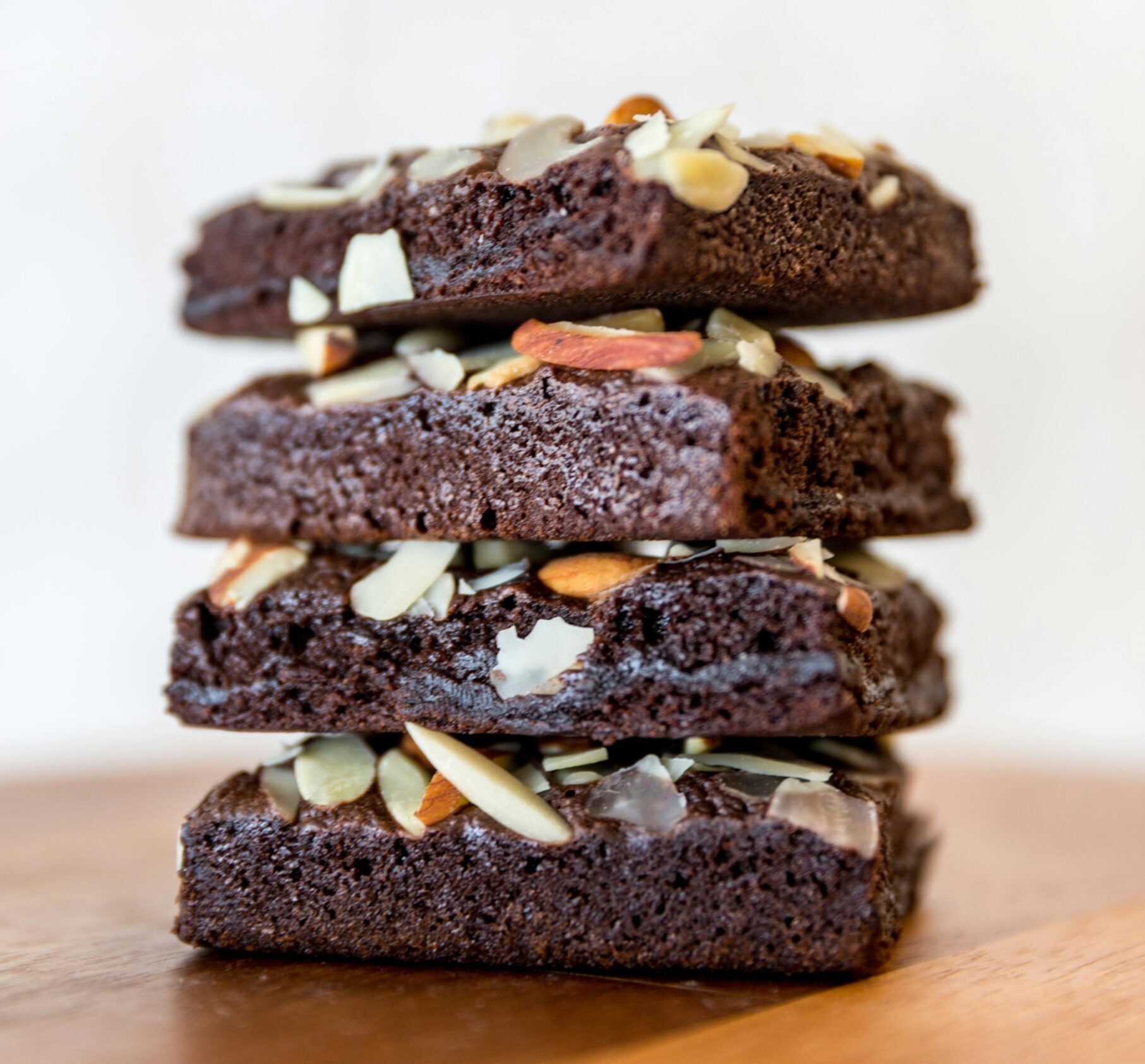 Close-up of stacked chocolate brownies topped with almond slices on wooden surface.